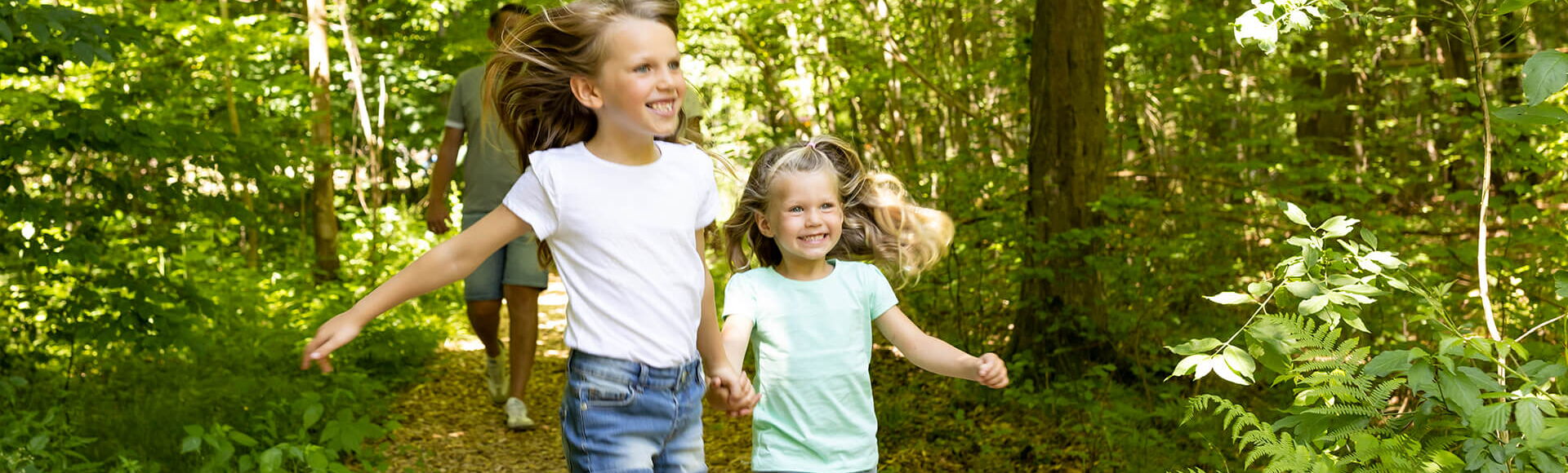 Zwei Mädchen laufen freudig auf einem Pfad durch den Wald, im Hintergrund der Vater.