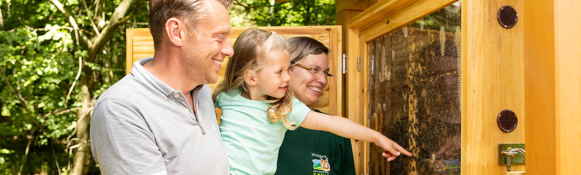 Frau Engelbrecht steht gemeinsam mit Vater und Tochter vor einem Bienenstock im Fensterkasten, Vater hält Tochter auf seinen Armen, während die Tochter auf den Bienenstock zeigt