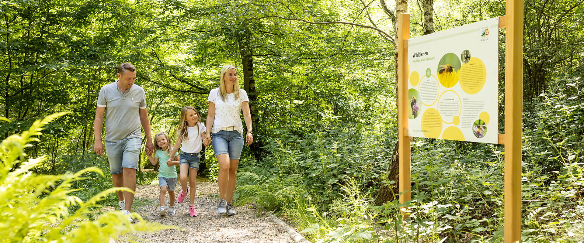 Eltern spazieren mit Ihren beiden kleinen Töchtern durch den Bienenwald des Wildparks Knüll und betrachten das Infoschild.